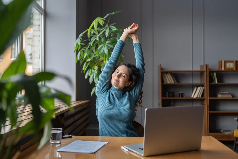 A woman stretches her arms while sitting at a desk in front of her laptop, promoting the importance of taking breaks to relieve tension and support spinal health, a key focus in chiropractic care - Chiropractor Louisville KY