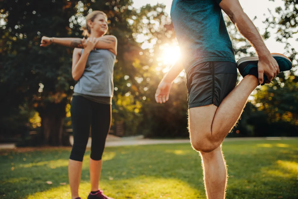 A man and woman stretch their arms and legs outdoors in a park, highlighting the importance of flexibility and mobility for joint and spinal health, often emphasized in chiropractic care - Chiropractor Louisville KY